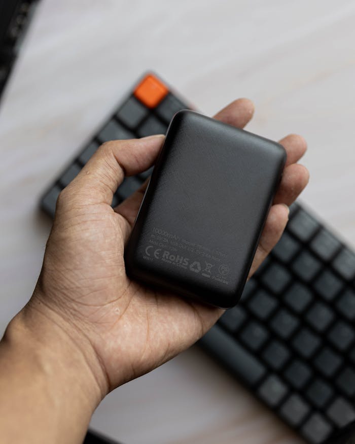 A close-up shot of a hand holding a black power bank with a keyboard in the background.